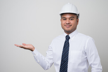 Young asian civil engineer showing palm up hand to blank space. Worker wearing hard hat standing on isolated white background.