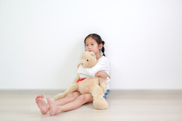 Asian little child girl hugging a teddy bear doll sitting against white wall in the room.