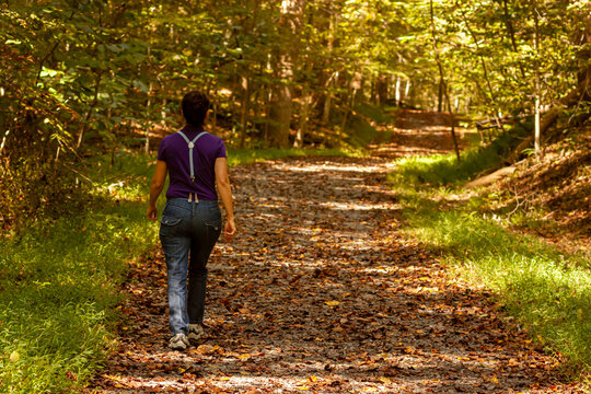 A woman wearing jeans with suspenders is walking alone in a hiking trail covered with fallen autumn leaves. The trail goes through a forest and is shaded. A fall concept image with some mystery.