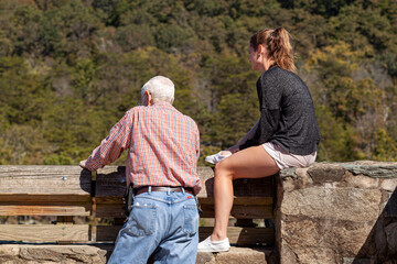 Obraz na płótnie Canvas An elderly man and his teen daughter are looking at the great falls, from a scenic overlook. The girl wearing shorts is sitting on stone wall despite the risk of fall