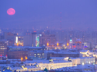 Fototapeta premium Big red full moon over Omsk, full moon over the city center. Lenin street, Assumption Cathedral and a smoking oil factory against the backdrop of the city with the setting moon in the morning.