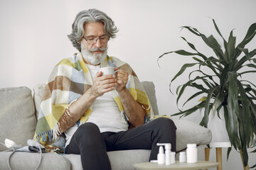 Senior man alone sitting on sofa. Sick man covered with plaid. Grangfather with cup of tea.