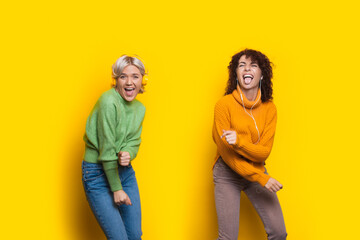Crazy women with headphones are dancing on a yellow studio wall wearing casual clothes and smiling at camera