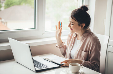 Lovely young woman greeting somebody on laptop while reading a book and drinking a coffee sitting...