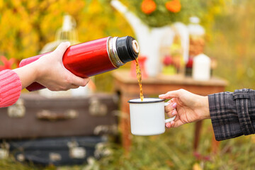 Cup of tea or coffee.  Pour hot tea or coffee from a red thermos into a mug.  Fragments of two girls' hands. Autumn background