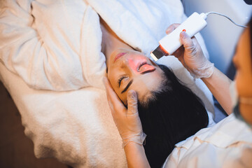 Upper view photo of a young lady lying in bed and doing facial procedures with apparatus at the spa salon