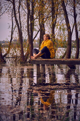 A young girl in a yellow sweater sits on the bridge over the lake and catches fish.