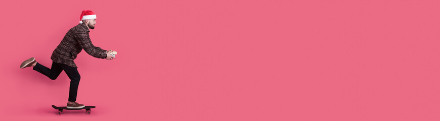 Skating caucasian man with a present and red santa hat is advertising something on a studio wall with free space