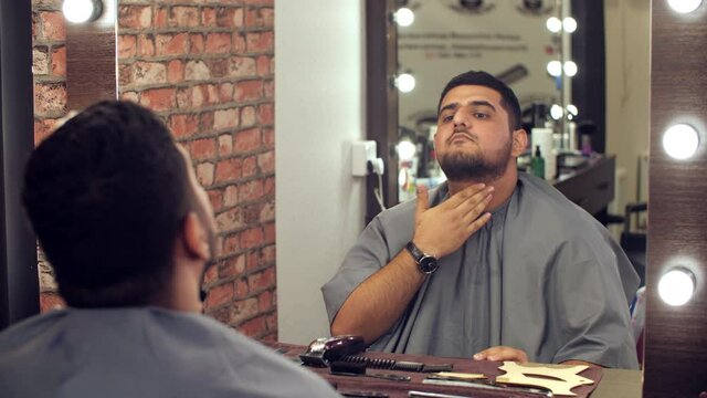Handsome man touching beard and looking at mirror in barber shop. Reflection of man in mantle sitting and checking his facial hair after grooming and styling in barbershop