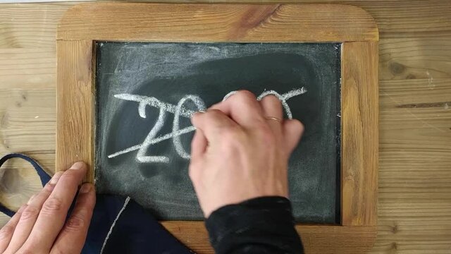 Male hand irritable crosses out year 2020 with piece of chalk on chalk plate in wooden frame