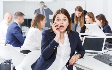 Obraz premium Smiling young female emotionally talking on mobile in busy modern open plan office