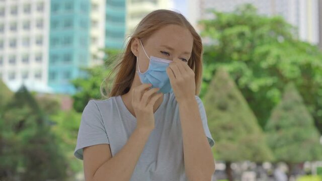 Young woman on a street wearing medical face mask in order to protect herself against Covid-19. Woman acts in a wrong way, she touches and scratches her face and mask