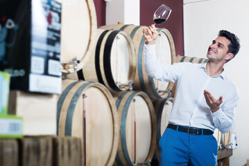 Cheerful man holding glass of red wine in shop with woods and tasting it