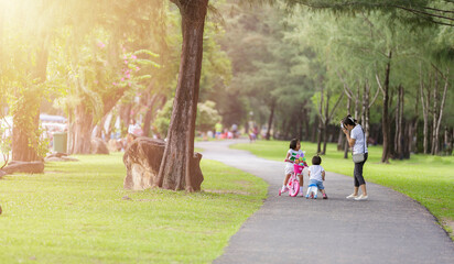 Fototapeta premium Family in park concept. Mother and her daughter play at park together. Mother shot her daugther with camera. Little girls relax with bicycle on road in park. Young mother shot picture her daughter