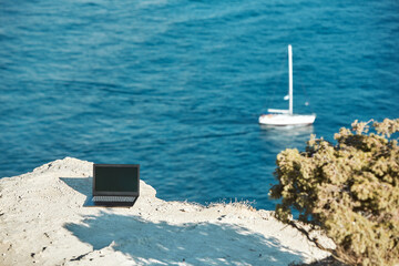 Fototapeta na wymiar Laptop on sea rock. Sea view and yacht on background. Distance work and freelance job concept. Travelling and work idea