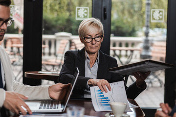 Portrait of senior businesswoman looking at camera while sitting in cafeteria with her colleagues.