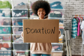 African american girl holding donation box and smiling at camera, posing in front of boxes full of clothes, Young volunteer working for a charity