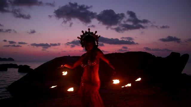 Erotic and sensual belly dancer, performing spinning moves with fire palm torch props, on the beach during golden hour. Slow motion.