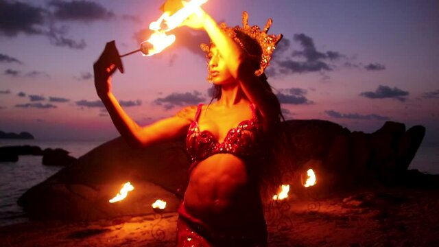 Erotic and sensual belly dancer, performing with fire palm torch props, on the beach during golden hour. Slow motion.