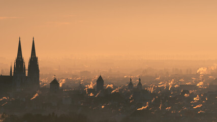 Overview of city skyline at the beginning of the heating period in autumn with many smoking chimneys in morning backlight twilight in Regensburg, Bavaria, Germany - obrazy, fototapety, plakaty