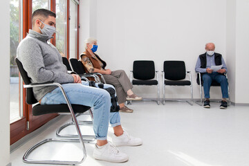 Young and old people with face masks keeping social distance in a waiting room of a hospital or...