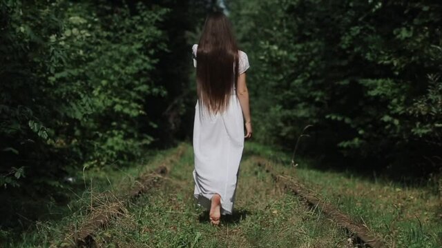A young woman in a white dress and with long hair walks barefoot on the grass between the old rusty rails of the railway. Back view. Close-up. The camera moves from bottom to top