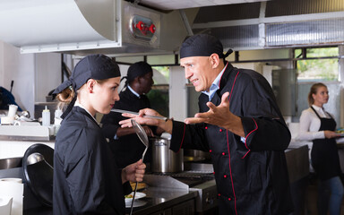 Fototapeta na wymiar Enraged chef expressing dissatisfaction with work of frustrated girl in restaurant kitchen