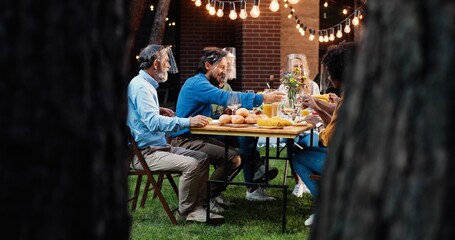 View over trees on mixed-races cheerful people in masks shields talking and eating at picnic. Multi ethnic family having picnic in back court and chatting. Communication during pandemic.