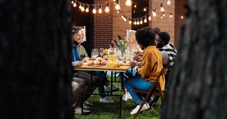 View over trees on mixed-races cheerful people in masks shields talking and eating at picnic. Multi ethnic family having picnic in back court and chatting. Communication during pandemic.