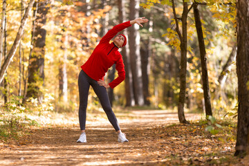 Warm up before jogging. Caucasian adult woman in red sportswear and a cap does side bends in the forest in autumn outdoor, selective focus