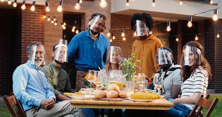 Portrait of mixed-races people in face shields at dinner table in back yard and smiling to camera. Multi ethnic family at picnic in court outdoors. Pandemic communication of generations.