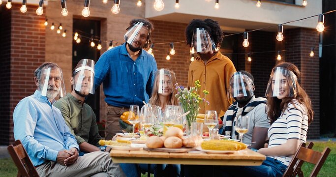Portrait of mixed-races people in face shields at dinner table in back yard and smiling to camera. Multi ethnic family at picnic in court outdoors. Pandemic communication of generations.