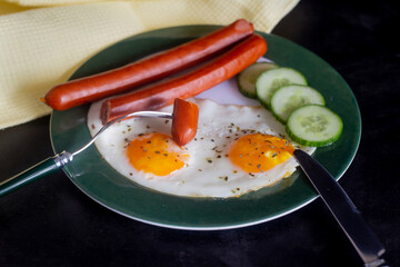 Breakfast with scrambled eggs, sausages and cucumber. On a dark background with a yellow napkin