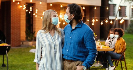 Portrait of Caucasian middle-aged couple in medical masks hugging in yard at picnic. Man and woman in hugs at court barbeque looking at each other with love and smiling to camera.