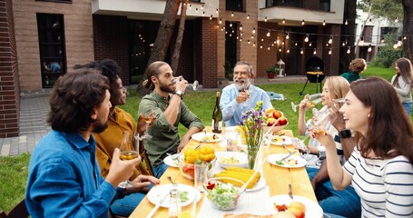 Happy multi ethnic family sitting at table with meal outdoor at picnic and toasting with juice and wine. Joyful young and old people having dinner and toast at party barbrque. Celebration on weekend.