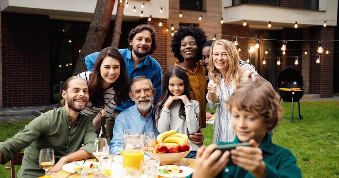 Mixed-races happy family at party dinner outdoor in yard smiling and posing to smartphone camera while small boy taking selfie photo. Multi ethnic people making photos together at barbeque Celebration