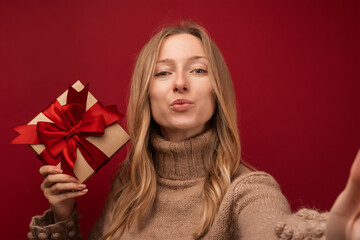 Charming blonde woman holding gift with red ribbon. Studio shot red background. New Year Birthday Holiday concept