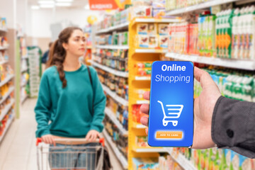 A person's hand holds a mobile phone, with a shopping basket on the screen. In the background, a woman is pushing a grocery cart in a supermarket, in a blur. The concept of online shopping