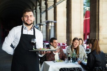 Cheerful waiter holding tray at restaurant with customers his behind