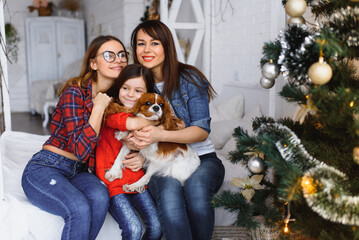 Two women and little girl with dog hugging near Christmas tree