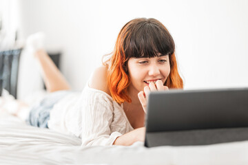 Young woman with colored hair lying on bed looking at a smart device while smiling