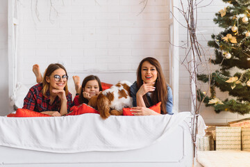 Two young beautiful women and a girl lie on the bed at home near the Christmas tree