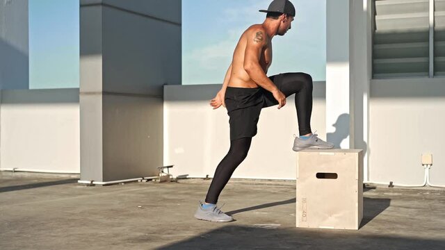 Fit shirtless athletic man jumping up to plyometric wood box outdoors on rooftop