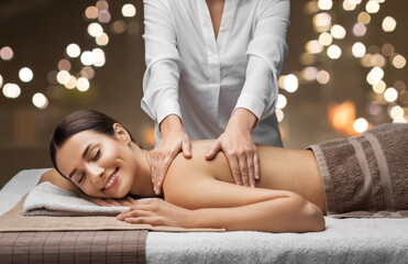 wellness, beauty and relaxation concept - beautiful young woman lying and having back massage at spa over festive lights on background