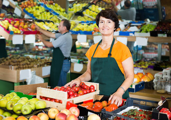 Positive female seller is standing with box with tomatos in the supermarket