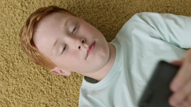 Flat lay of Caucasian teenage boy with ginger hair lying on carpet with mobile phone in his hands and having chat with friend by video connection