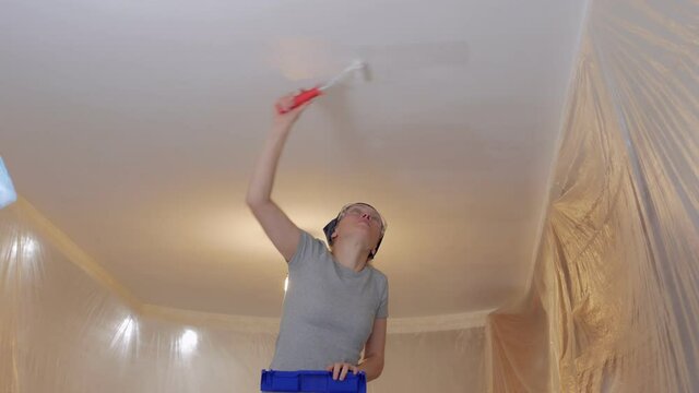 A woman standing on a ladder paints the ceiling white with a roller.