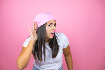 Young beautiful woman wearing pink headscarf over isolated pink background surprised with hand over ear listening an hearing to rumor or gossip