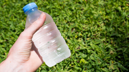 Hand holding blue plastic bottle of still water on the green grass. Summer day refreshment. Drink a lot of water