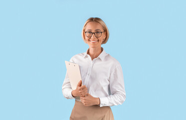 Portrait of happy businesswoman in formal wear and eyeglasses holding clipboard over blue studio...